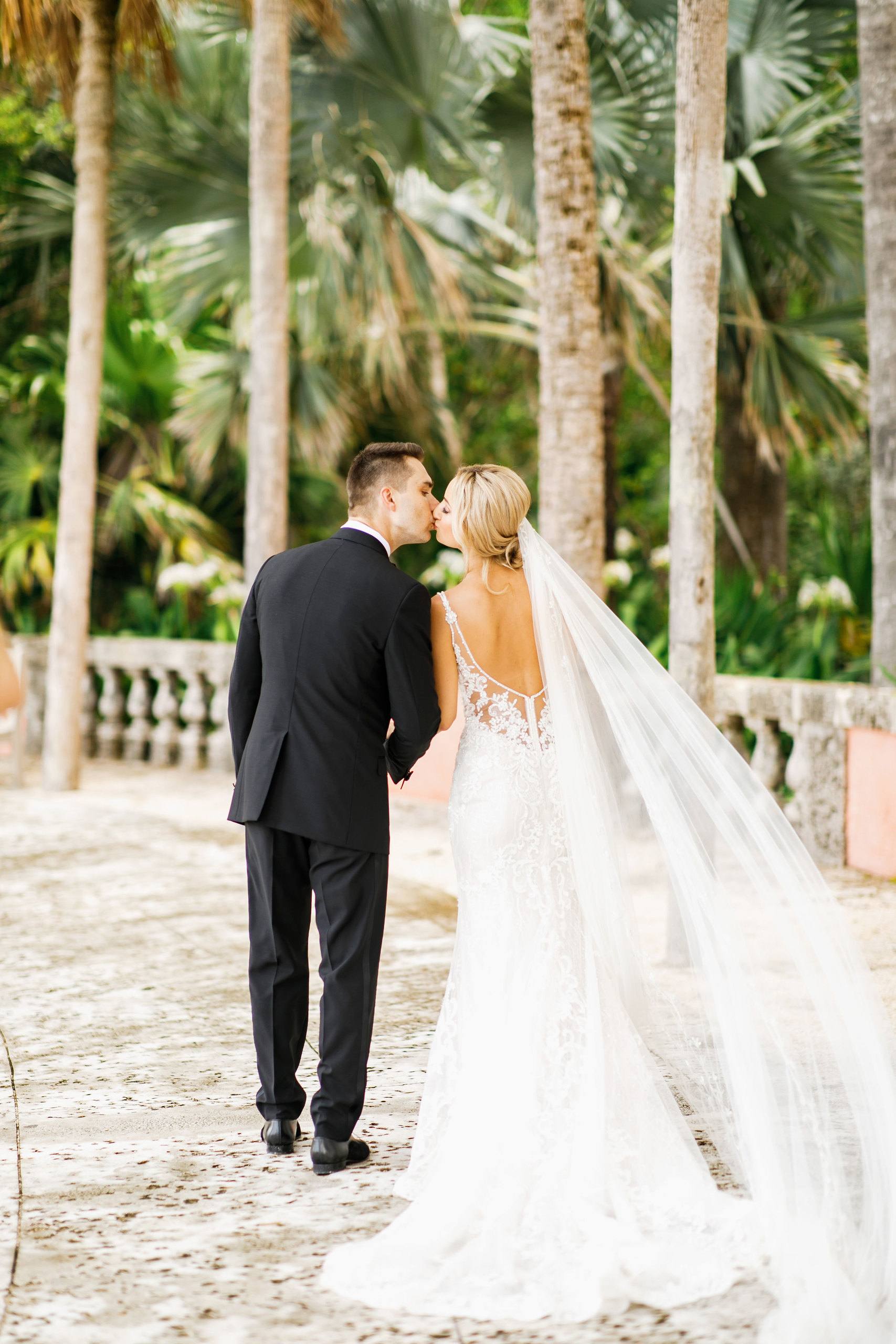 Bride and groom kissing under a long veil on a palm lined path