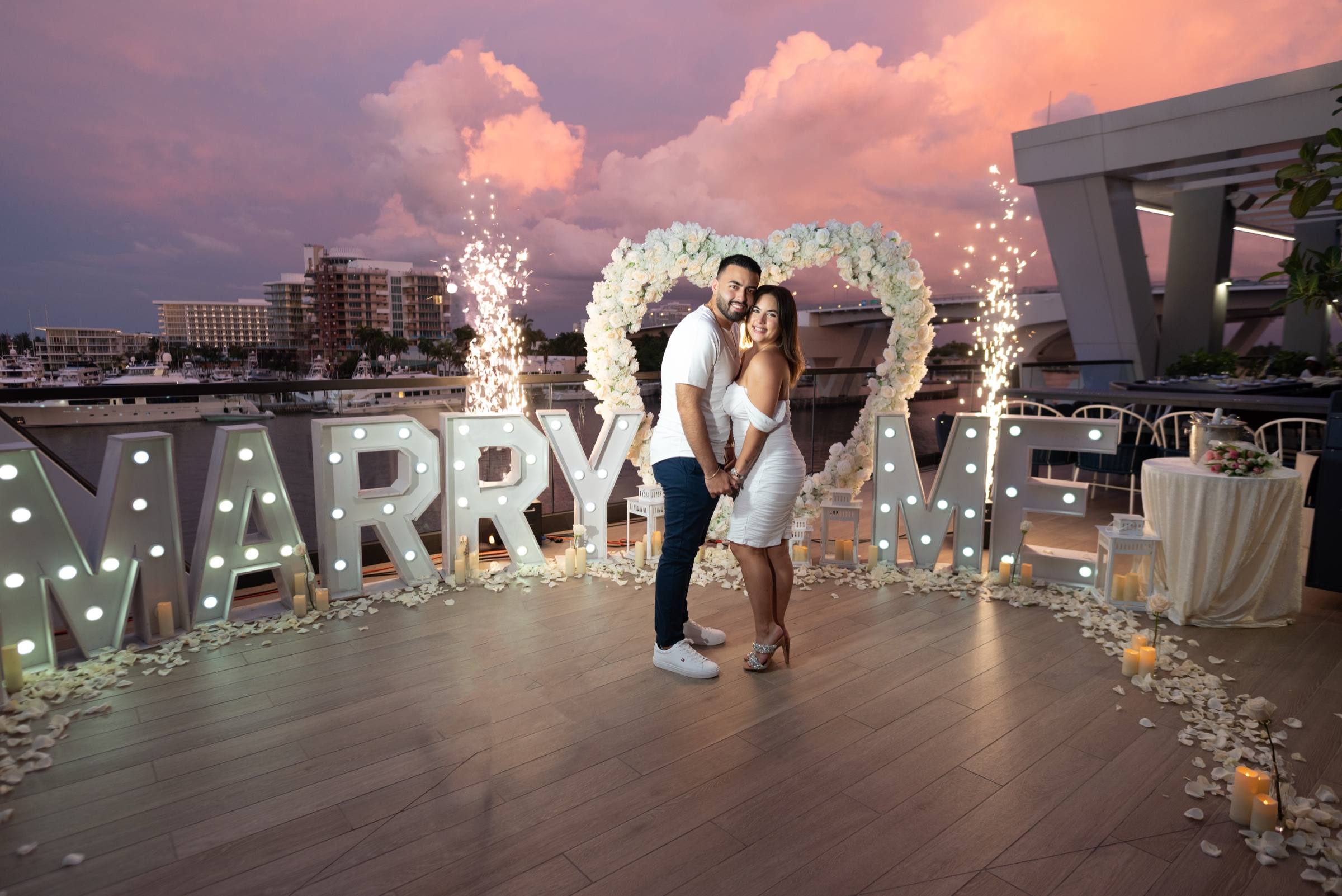 Illuminated Marry Me letters with a red floral heart, rose petals and candles on a private rooftop
