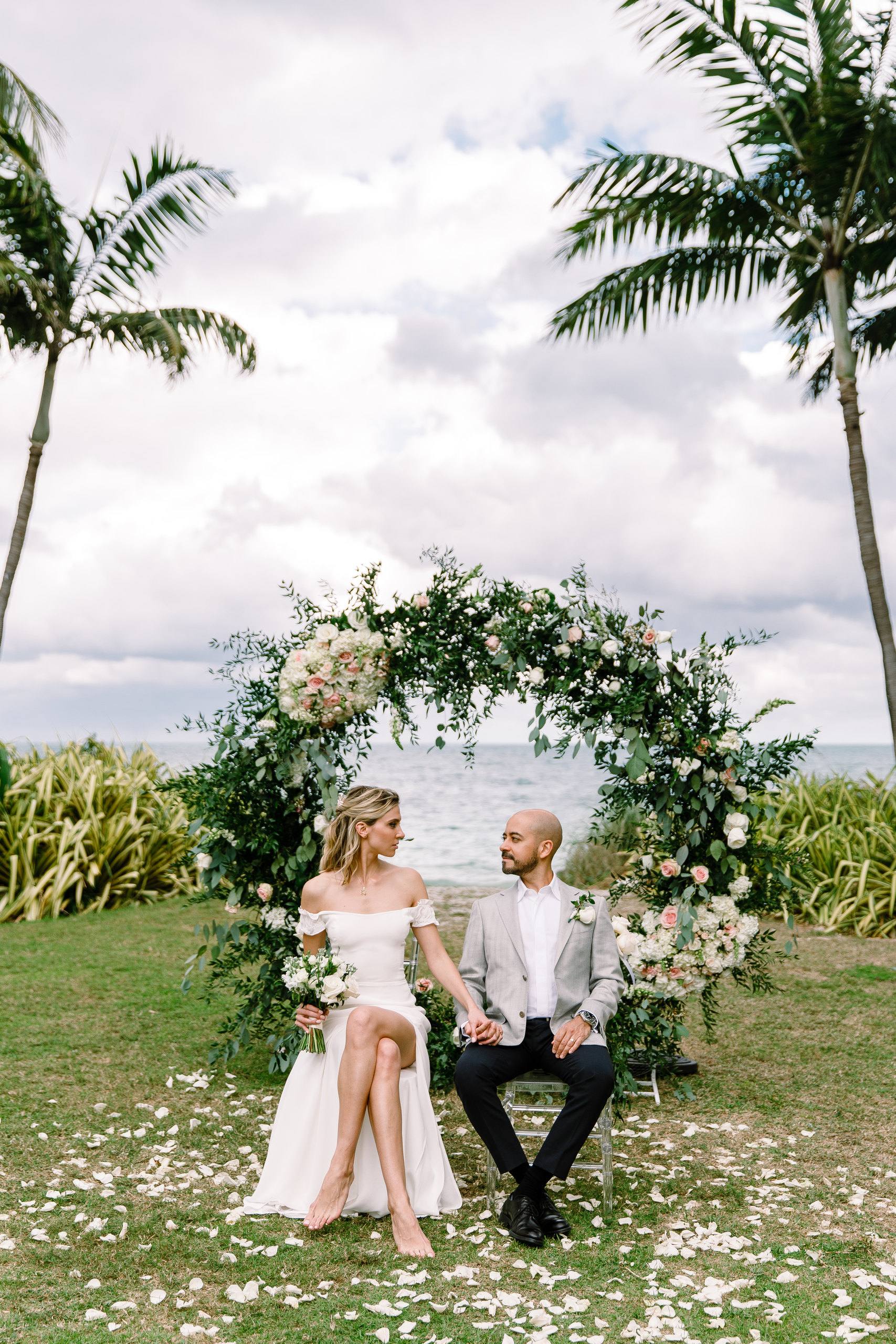 Couple seated under a circular floral arch on a lawn above the ocean