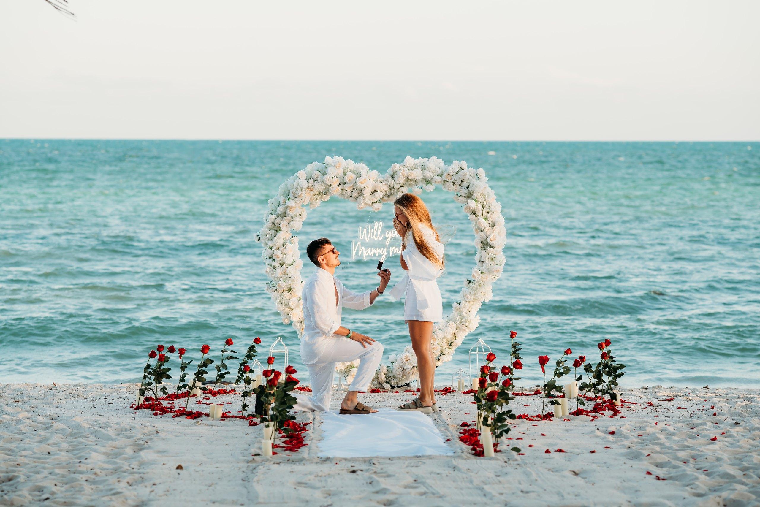 Man kneeling to propose under a white floral heart arch