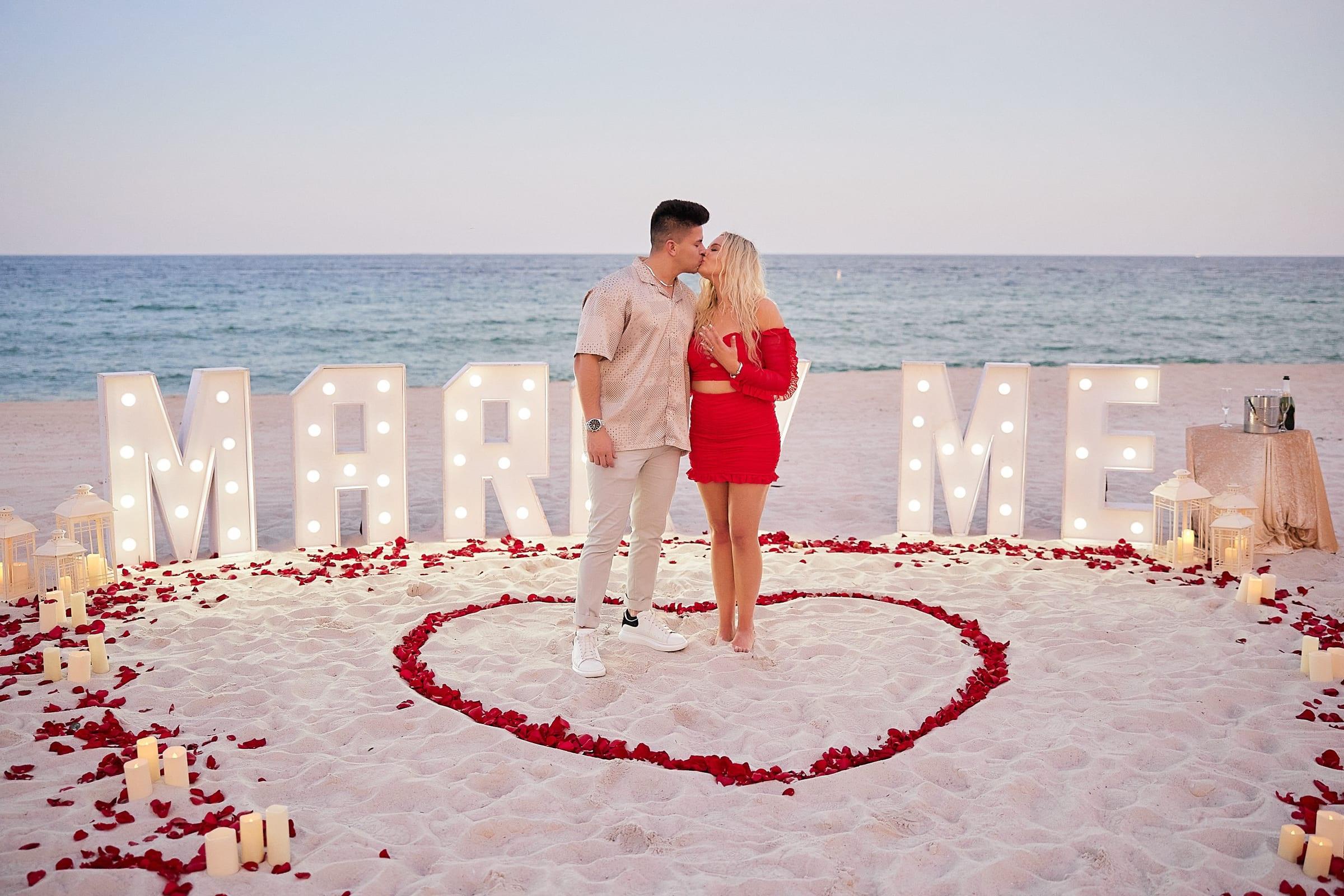 Couple kissing inside a heart of red rose petals in front of lit Marry Me letters on the beach