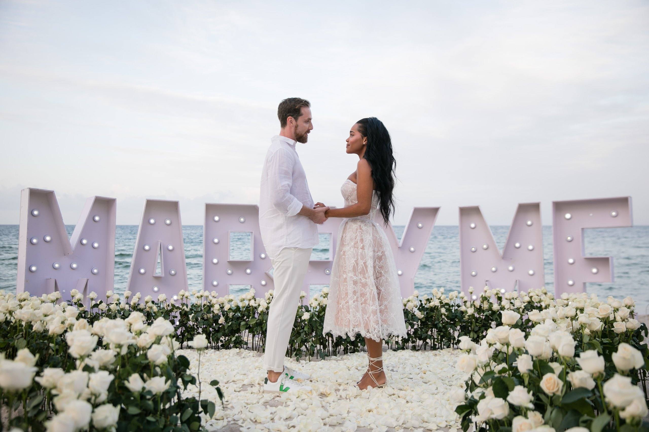Couple celebrating beside Marry Me letters and a heart of white roses