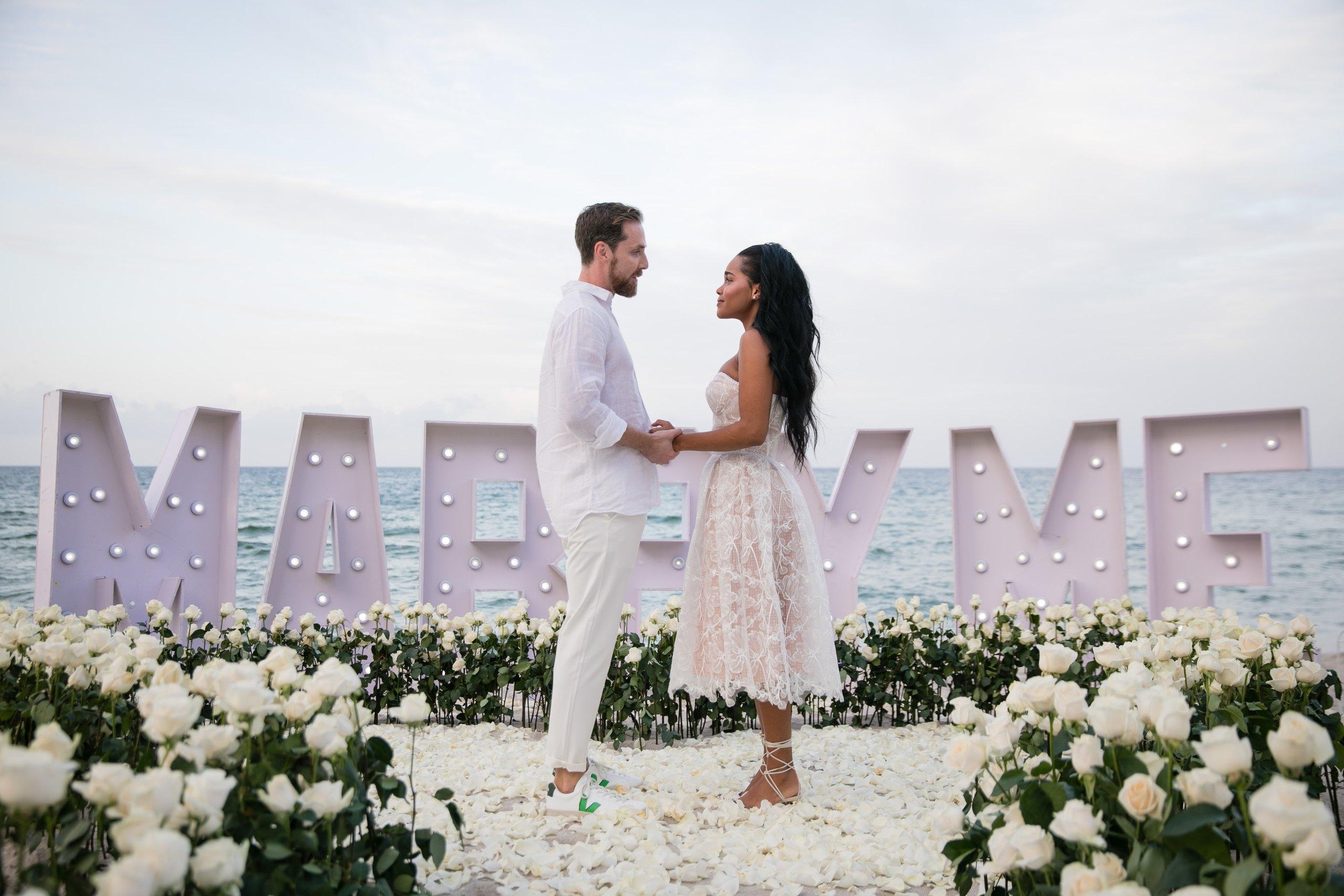 Couple celebrating beside Marry Me letters and a heart of white roses
