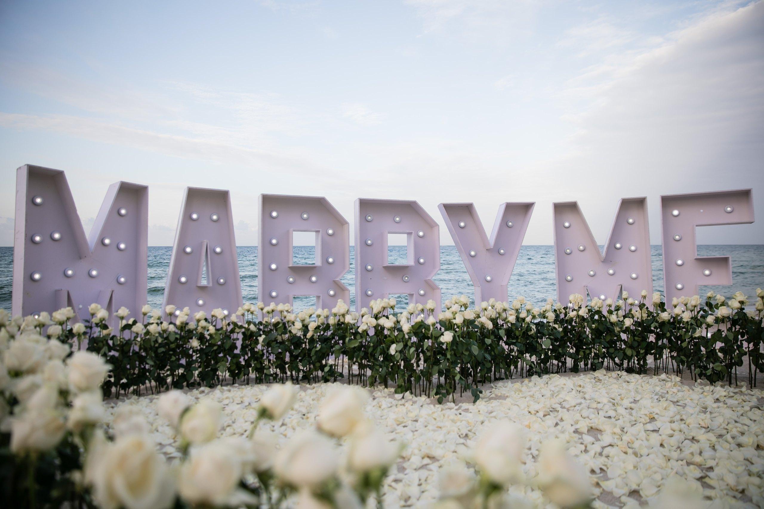 Large Marry Me letters framed by white roses and candelabras