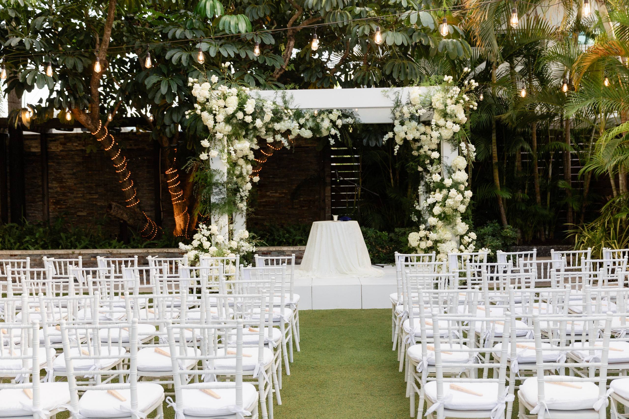 Rows of white chairs facing a floral arch in a garden courtyard